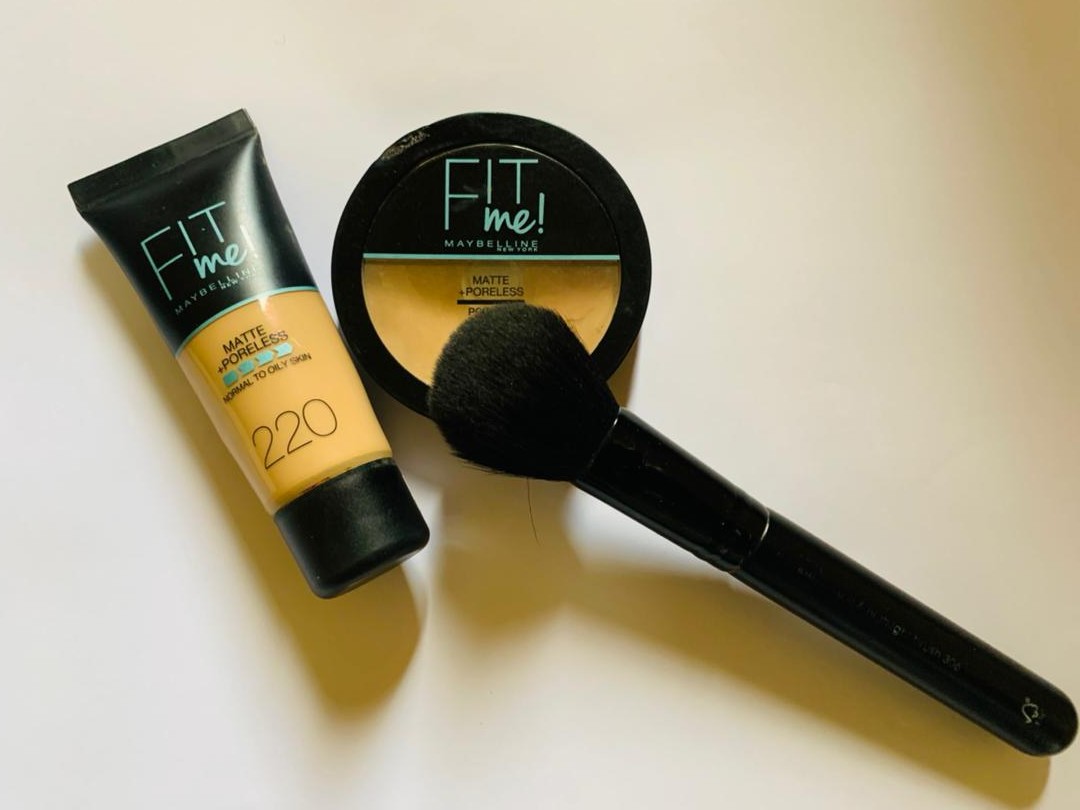 Produtos de maquiagem Maybelline Fit Me sobre uma superfície clara, incluindo base, pó compacto e pincel
