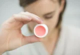Produto labial com cor em embalagem clara, segurado por uma pessoa.